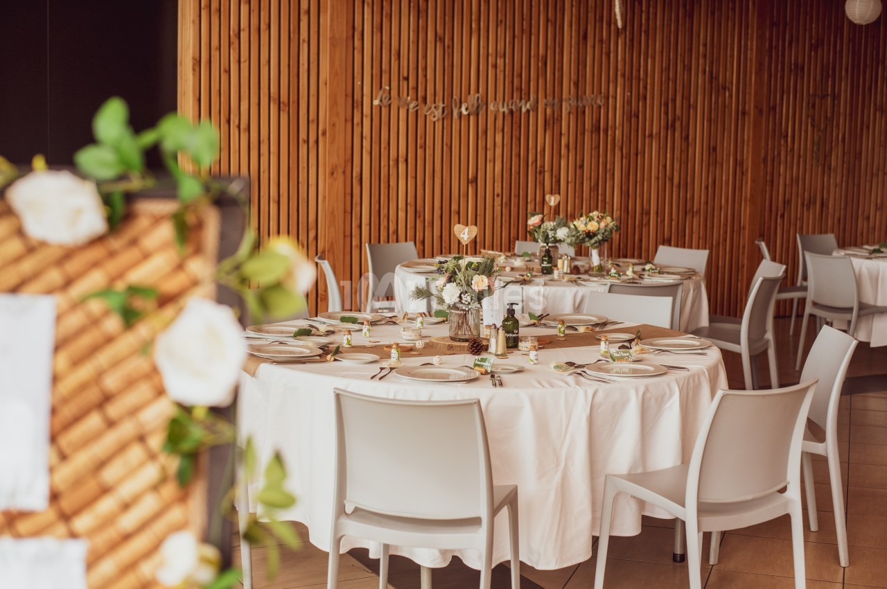 Salle décorée avec des tables rondes dressées, nappes blanches, fleurs et éléments de décoration élégants.