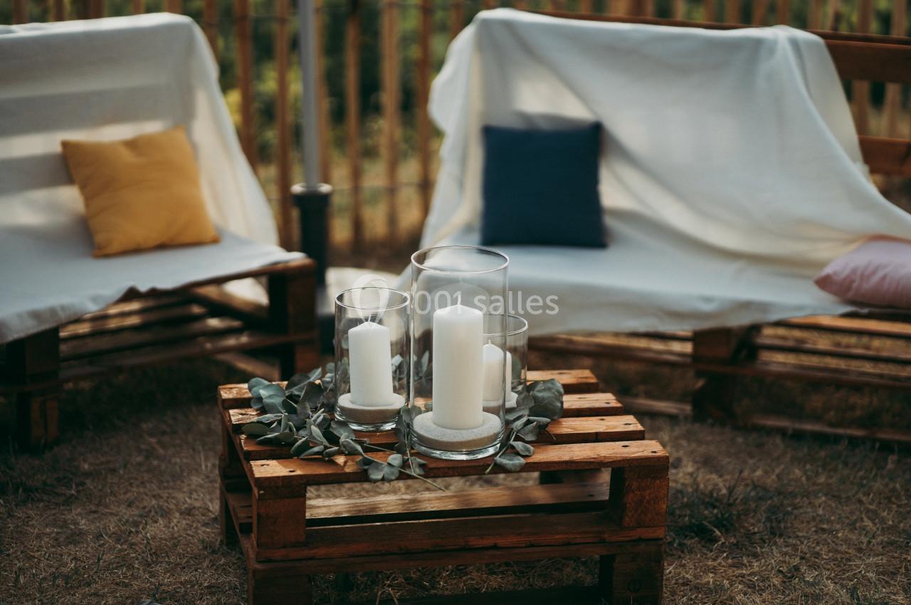Bougies blanches dans des photophores en verre sur une table en palettes, entourées de feuilles, avec des fauteuils en…