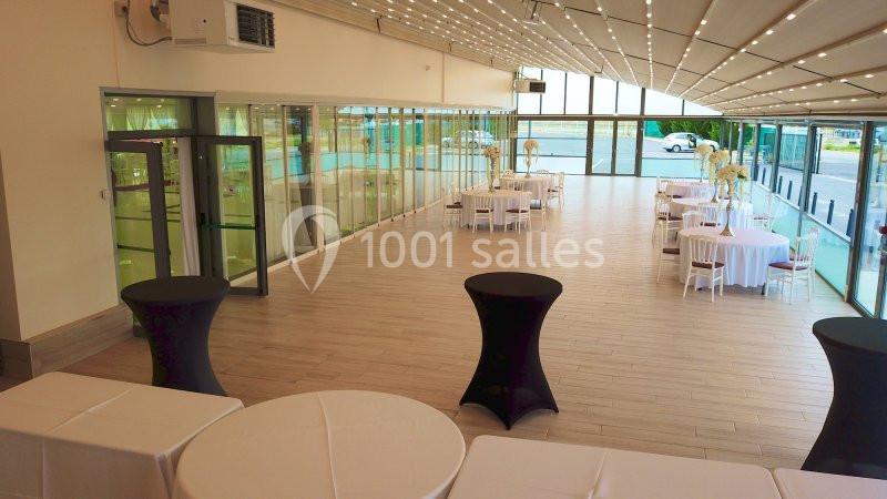 Salle lumineuse avec baies vitrées, tables rondes dressées et nappes blanches, vue sur un espace extérieur.