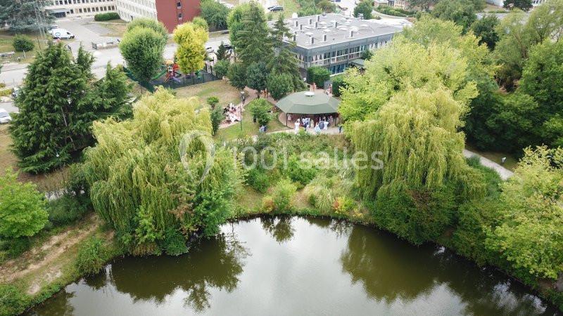 Vue aérienne d'un étang entouré de végétation dense, avec un bâtiment et une terrasse occupée en arrière-plan.