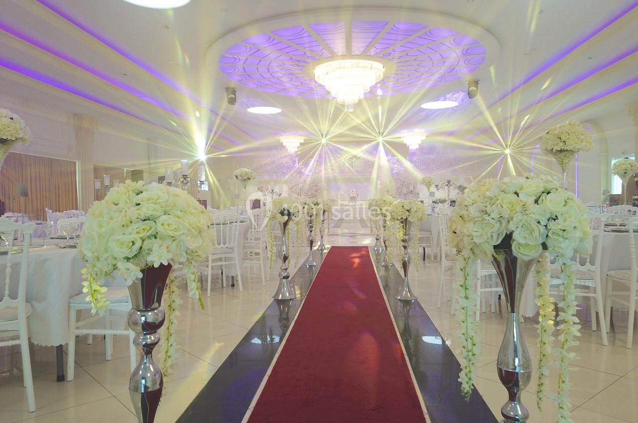 Salle de réception décorée avec des fleurs blanches, un tapis rouge central et des lustres illuminant le plafond.
