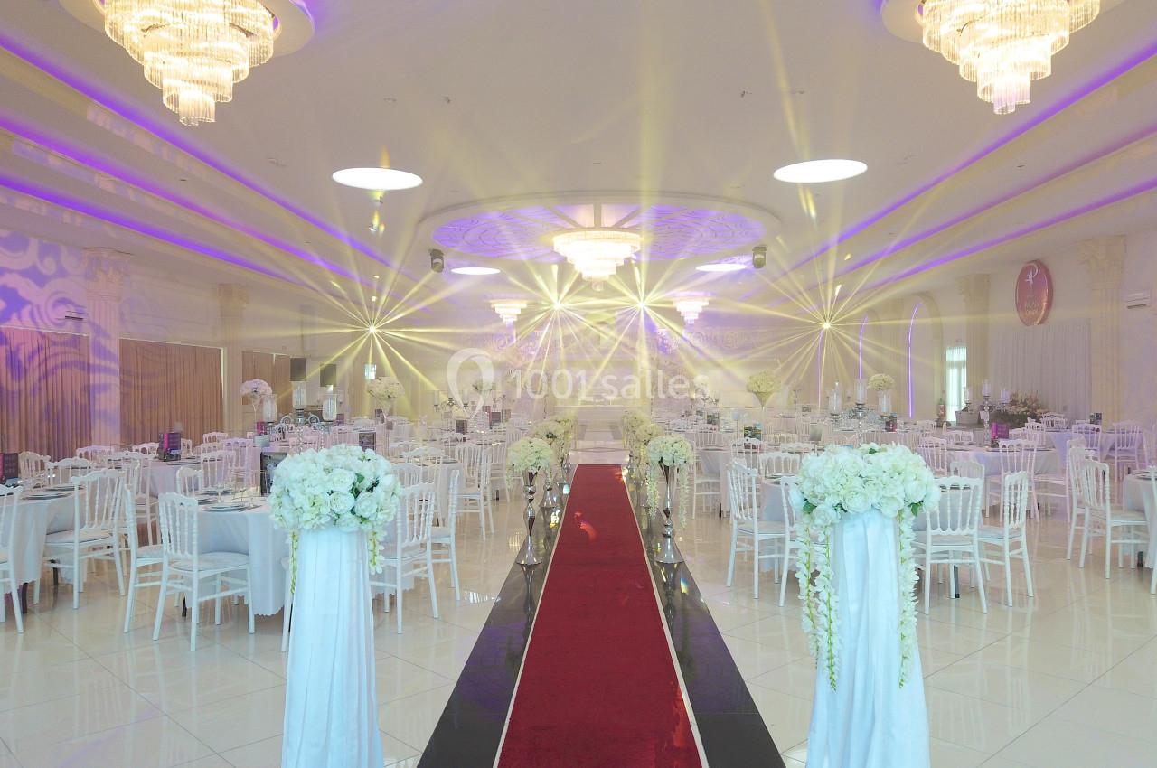 Salle de réception élégante avec tables décorées de fleurs blanches, tapis rouge central et lustres lumineux.