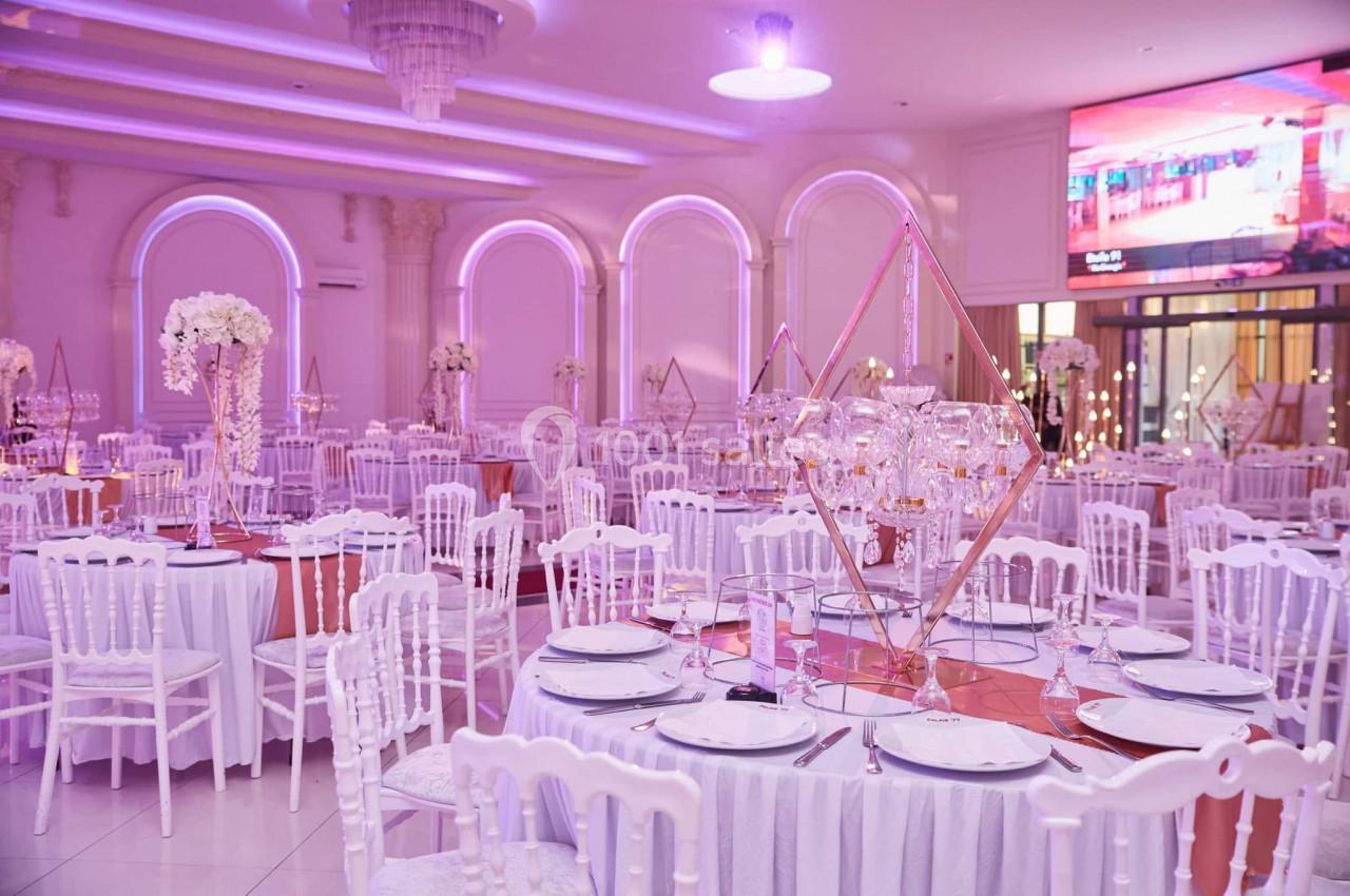 Salle de réception élégante avec tables décorées de nappes blanches, centres de table floraux et éclairage rose.