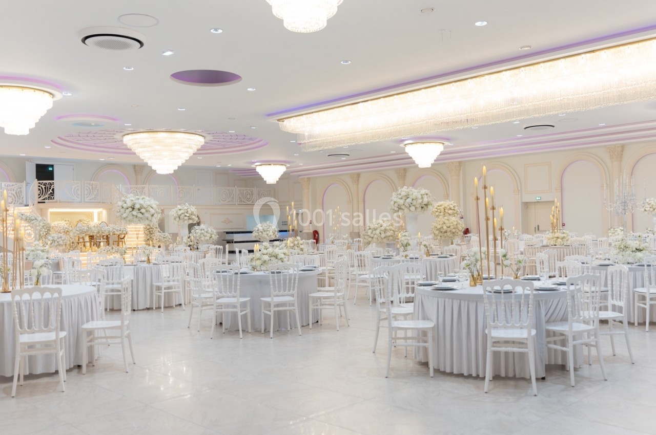 Salle de réception élégante avec tables rondes dressées, chaises blanches et décorations florales blanches.