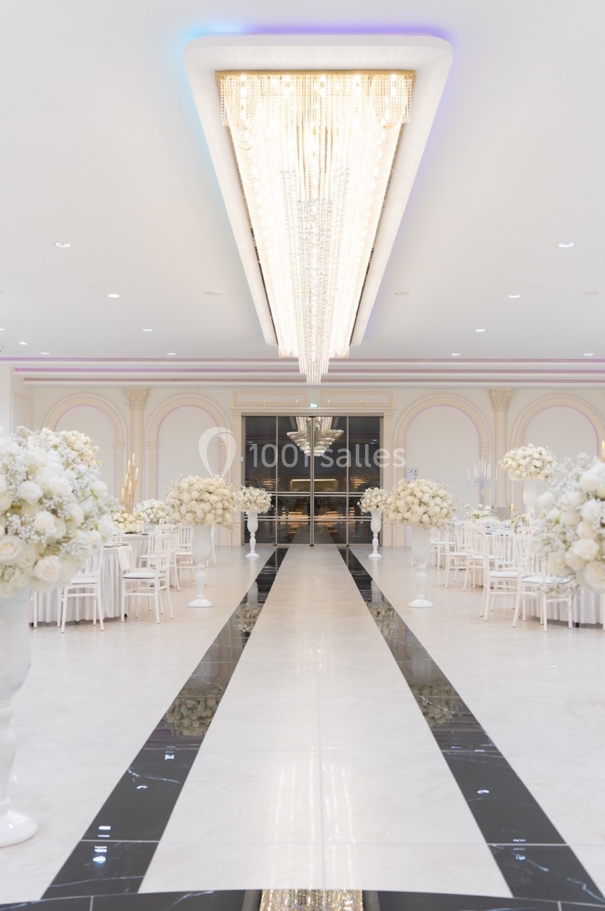 Salle de réception élégante avec un long lustre central, des tables décorées de fleurs blanches et un sol en marbre.