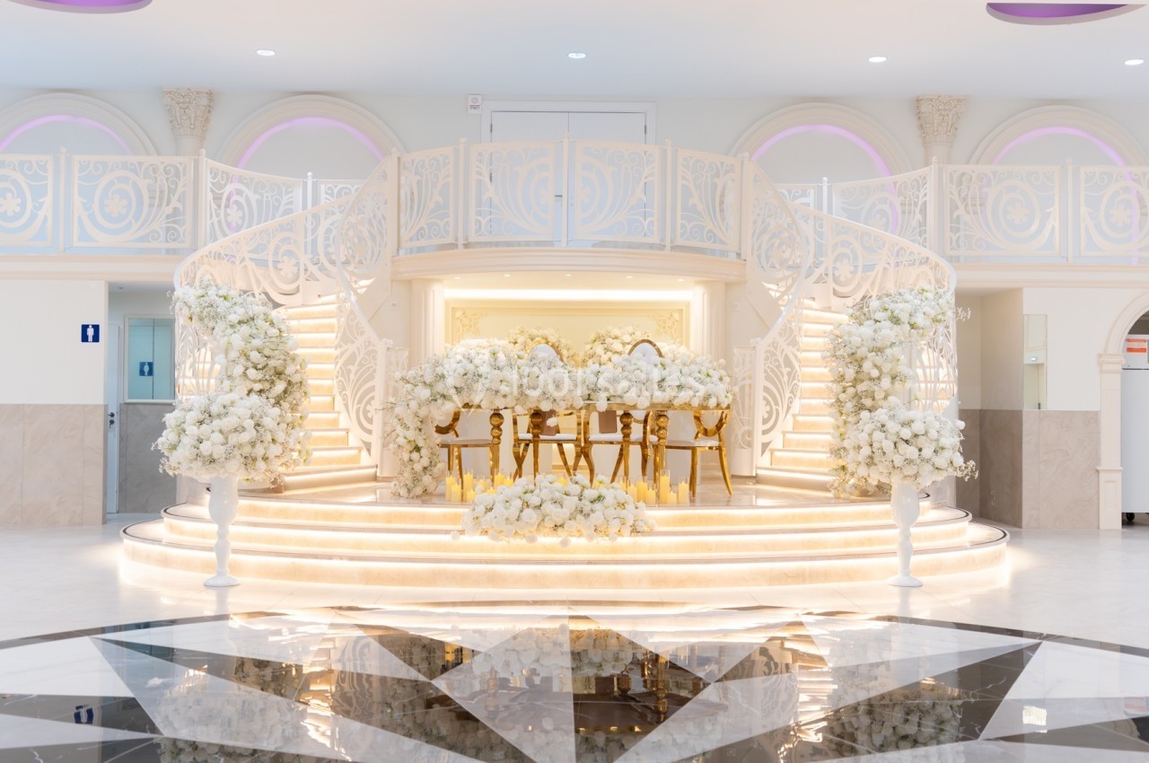 Escalier blanc en colimaçon entourant une scène décorée de fleurs blanches et de chaises dorées dans une salle lumineuse.