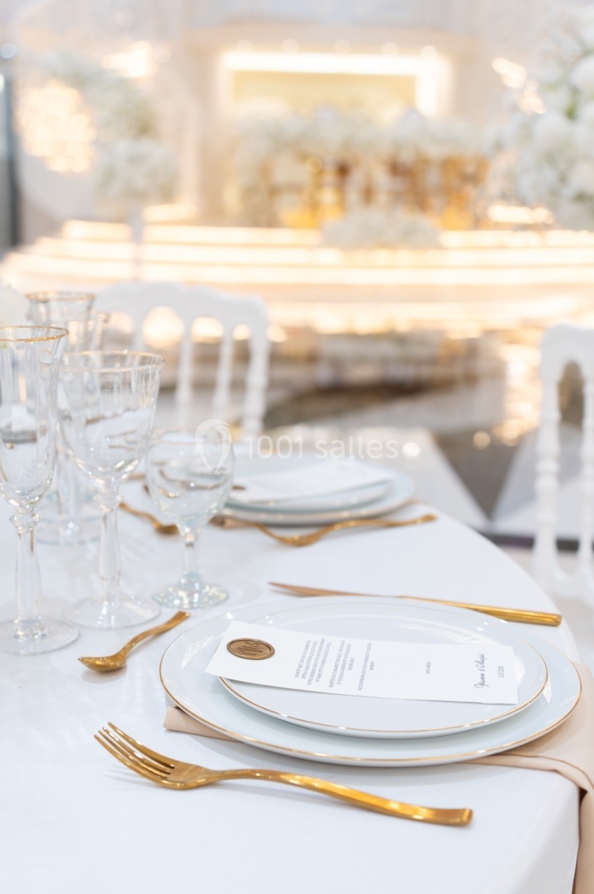 Table élégamment dressée avec vaisselle blanche, couverts dorés et menu, dans un décor lumineux et raffiné.