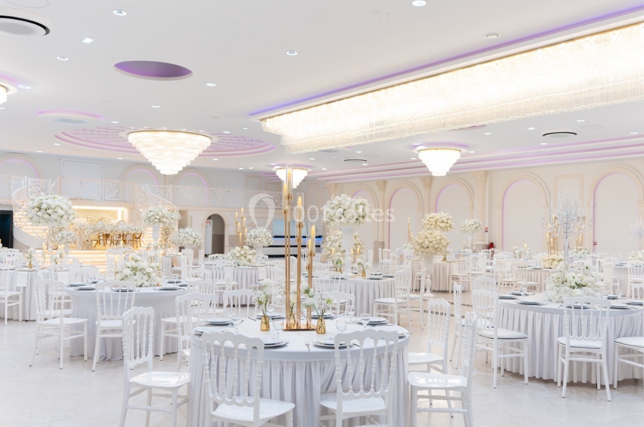 Salle de réception élégante avec tables rondes décorées de fleurs blanches et lustres imposants au plafond.