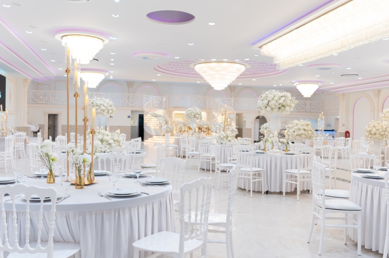 Salle de réception élégante avec tables rondes décorées de fleurs blanches, chandeliers dorés et lustres imposants.