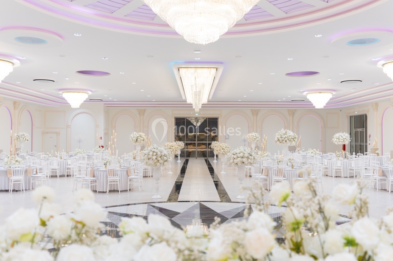 Salle de réception élégante avec tables décorées de fleurs blanches, lustres imposants et plafond orné.