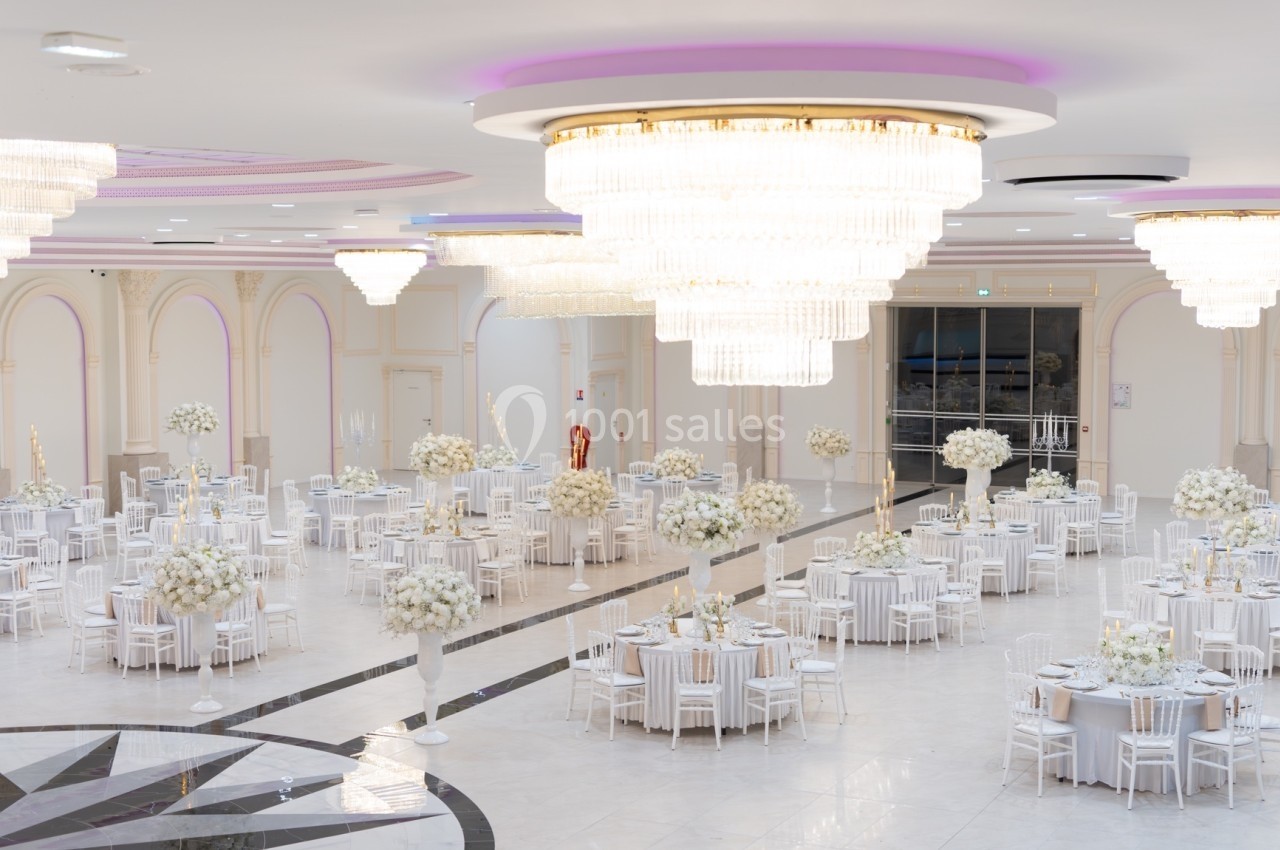 Salle de réception élégante avec tables rondes décorées de fleurs blanches et grands lustres suspendus.
