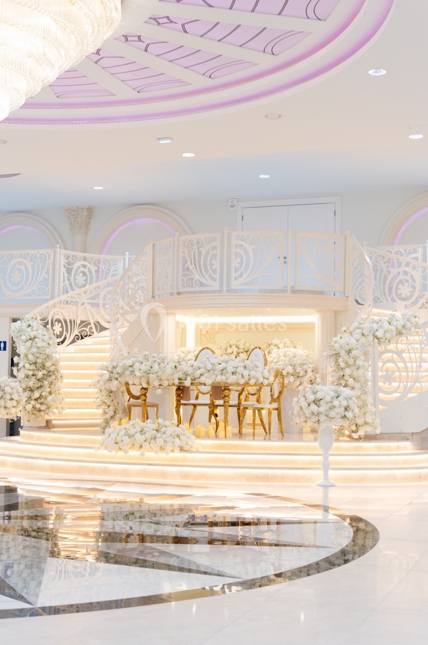 Salle de réception élégante avec escalier blanc, décor floral abondant et chaises dorées autour d'une table.
