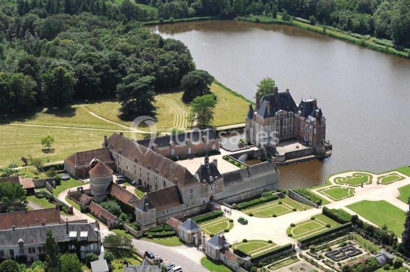 Vue aérienne d'un château entouré de douves, avec des jardins aménagés et un plan d'eau bordé d'arbres.