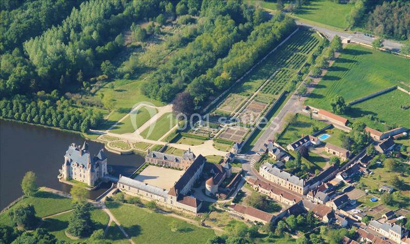 Vue aérienne d'un château entouré de jardins symétriques, d'un plan d'eau et de bâtiments annexes dans un cadre verdoyant.