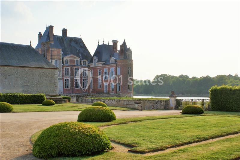 Château en briques et pierres bordant un jardin avec pelouses taillées et vue sur un plan d'eau en arrière-plan.