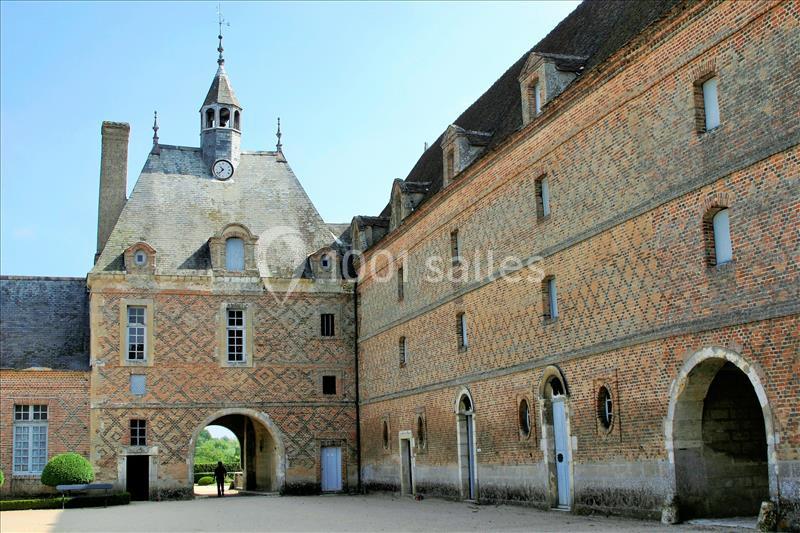 Bâtiment historique en briques avec une tour horloge, des arches et une cour pavée sous un ciel dégagé.