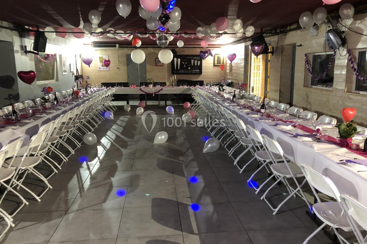 Salle décorée pour un événement avec des tables alignées, nappes roses, ballons suspendus et chaises blanches.