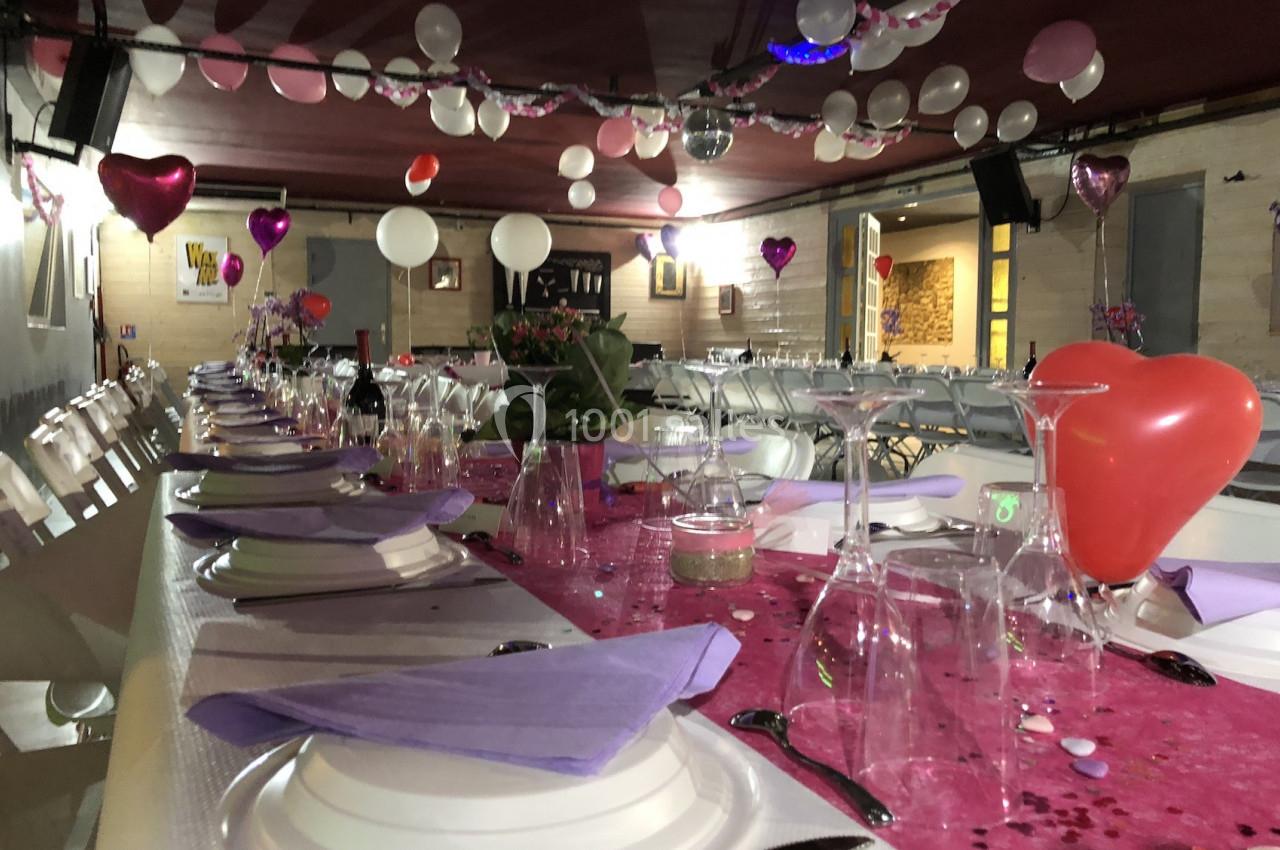 Salle décorée pour une fête avec des ballons suspendus, une table dressée avec nappes roses et serviettes violettes.