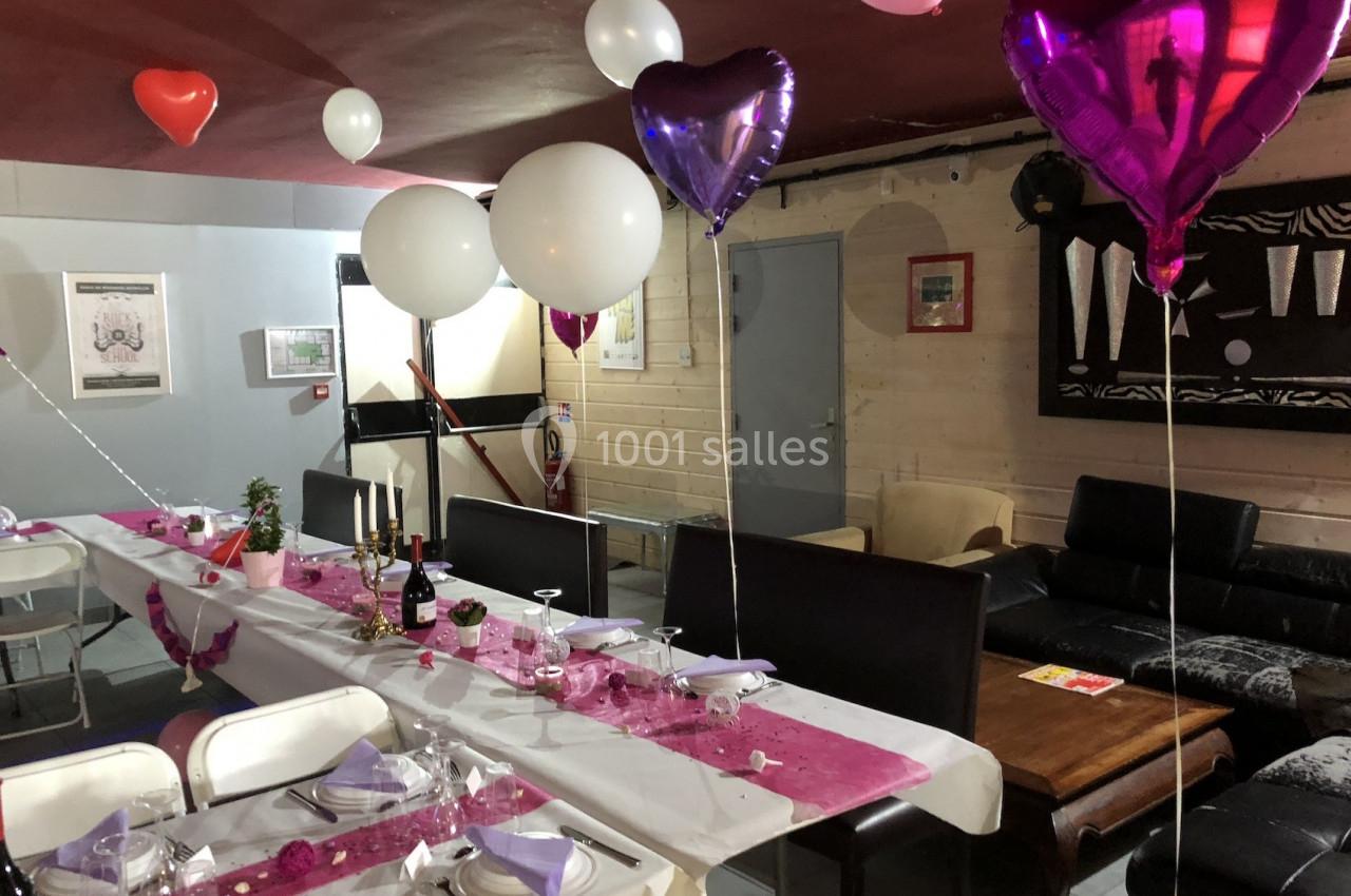 Salle décorée pour une fête avec des ballons en forme de cœur, une table dressée et des chaises blanches.