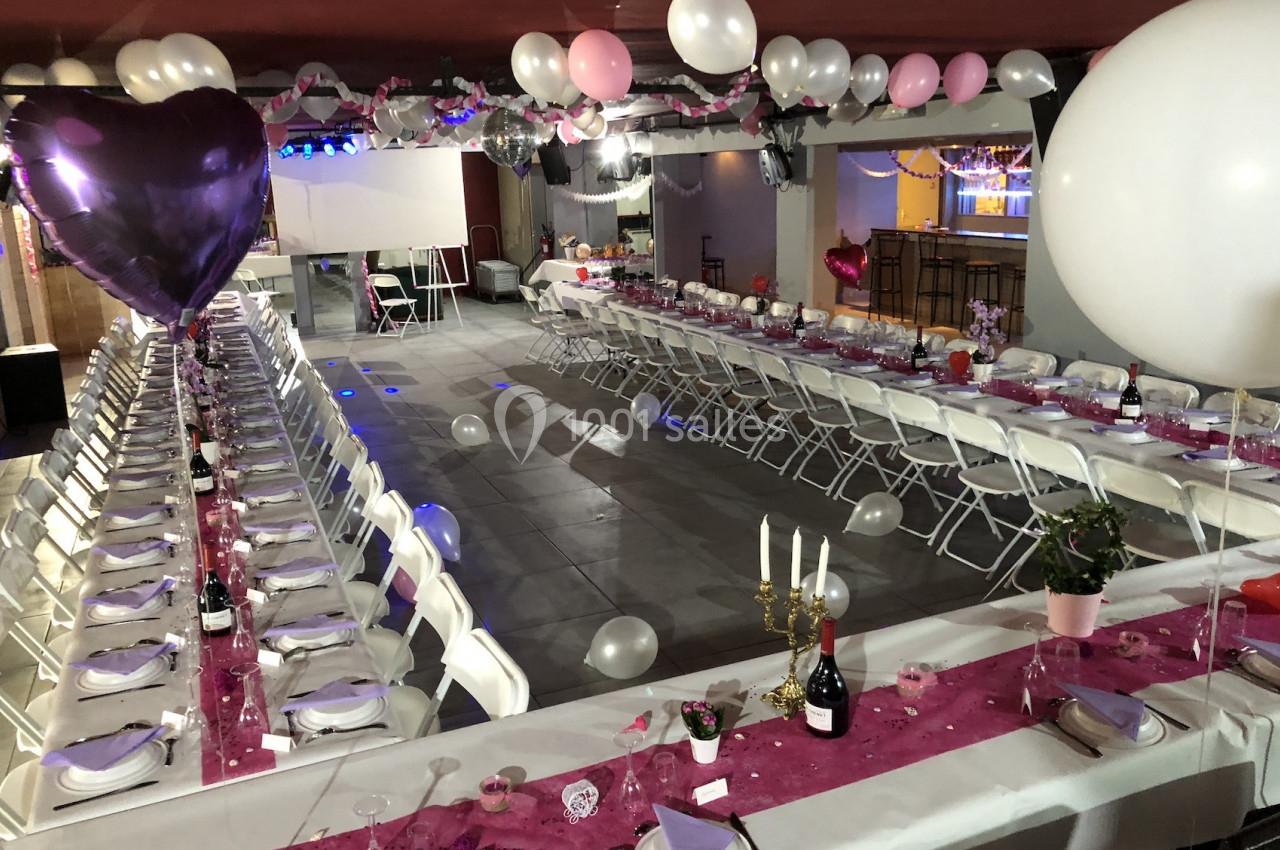 Salle décorée pour une réception avec longues tables dressées, nappes roses, ballons et chandeliers.