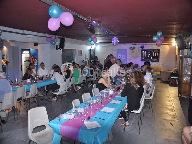 Salle décorée avec des ballons, des tables dressées et des convives assis lors d'un événement festif.