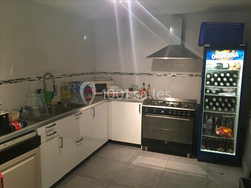 Cuisine équipée avec évier, cuisinière, micro-ondes, réfrigérateur vitré contenant des boissons et divers ustensiles.