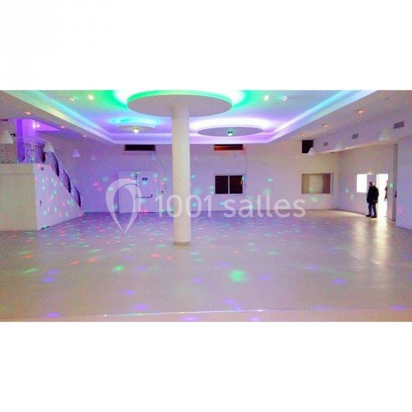Salle spacieuse avec éclairage coloré au plafond et reflets lumineux sur le sol, escalier visible à gauche.