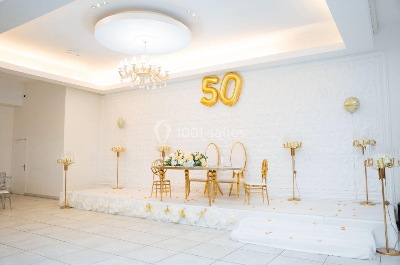 Salle décorée pour un 50e anniversaire, avec ballons dorés, table ornée de fleurs et chaises élégantes sur une estrade.