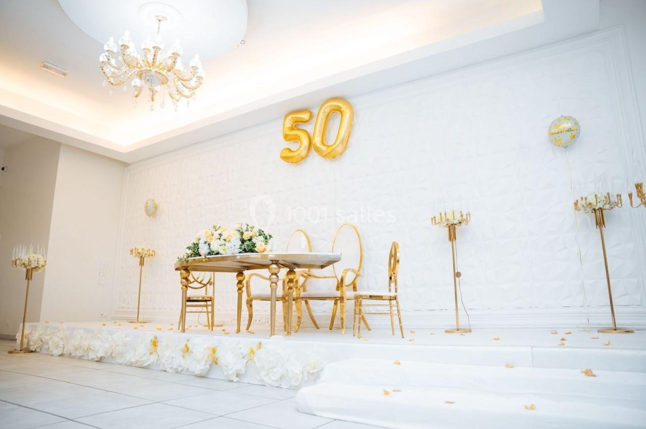 Salle décorée en blanc et or avec une table, des chaises dorées, des ballons ’50’ et des chandeliers élégants.