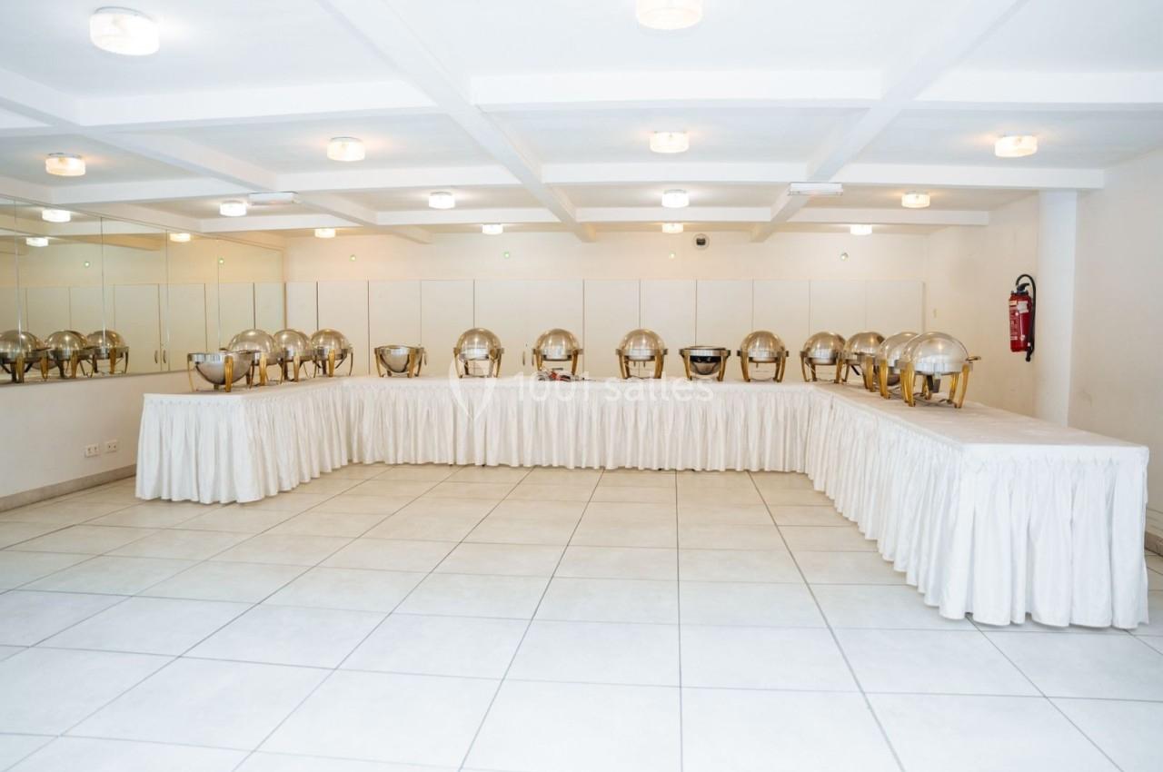 Salle lumineuse avec des plats en inox alignés sur des tables drapées de nappes blanches, prête pour un buffet.