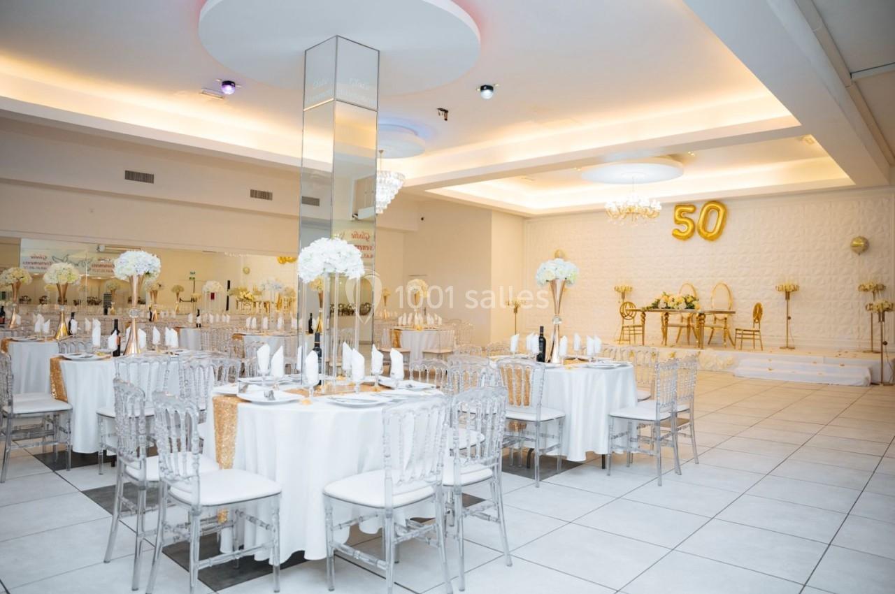Salle de réception élégante décorée en blanc et or, avec tables dressées et ballon ’50’ en arrière-plan.