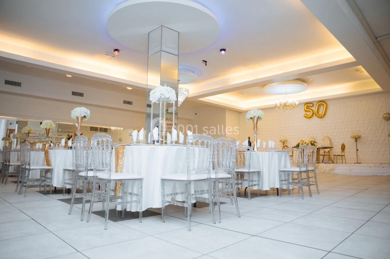 Salle de réception élégante avec tables rondes dressées, chaises transparentes et décoration florale blanche.