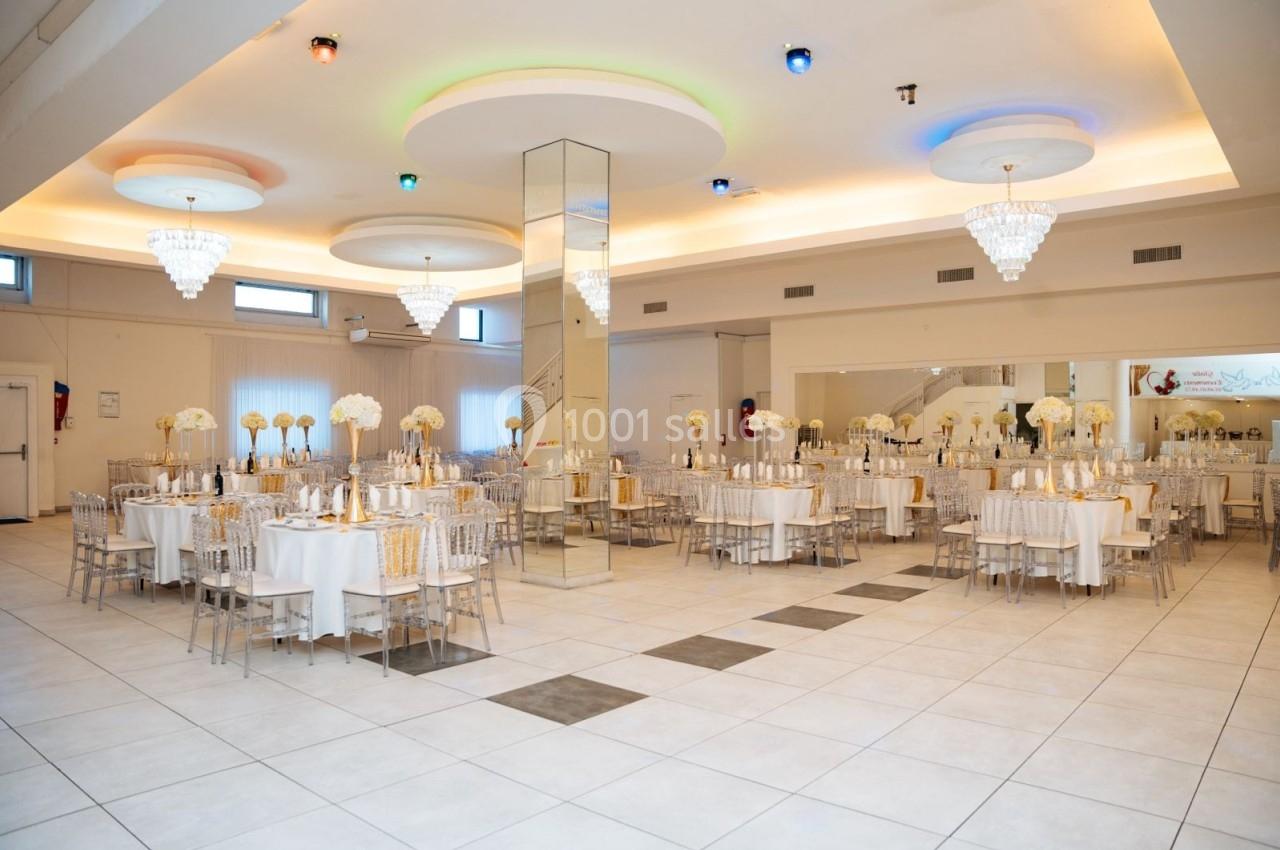 Salle de réception lumineuse avec tables décorées de fleurs blanches, chaises transparentes et lustres suspendus.