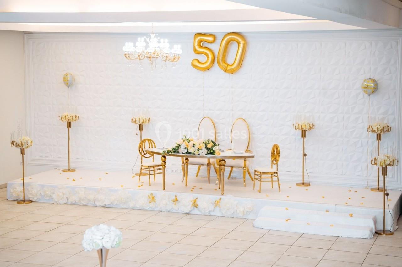 Salle décorée pour un 50e anniversaire avec ballons dorés, table ornée de fleurs et chaises élégantes sur une estrade…