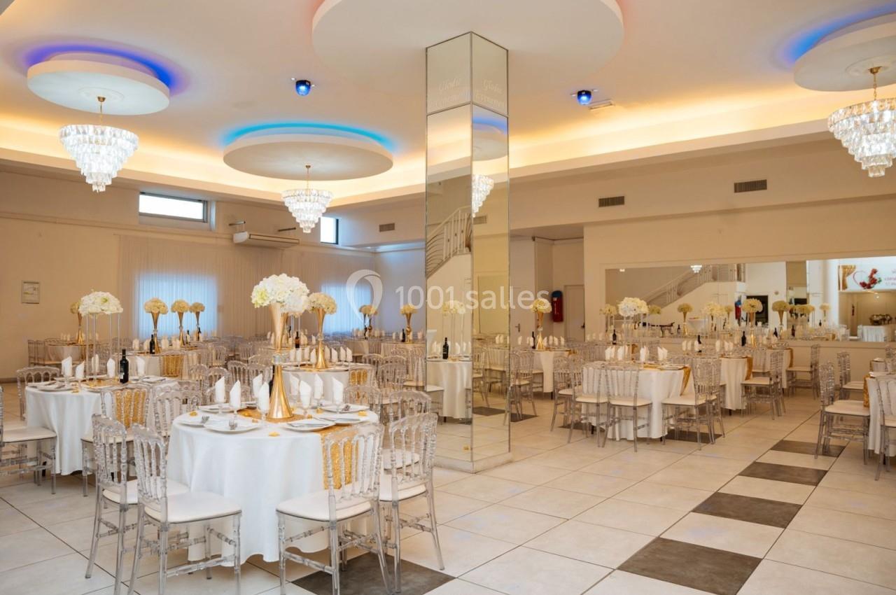 Salle de réception élégante avec tables rondes dressées, chaises transparentes et décorations florales blanches.
