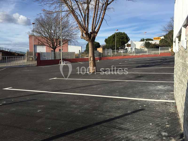 Parking vide avec marquages au sol, un arbre central et des bâtiments en arrière-plan sous un ciel dégagé.
