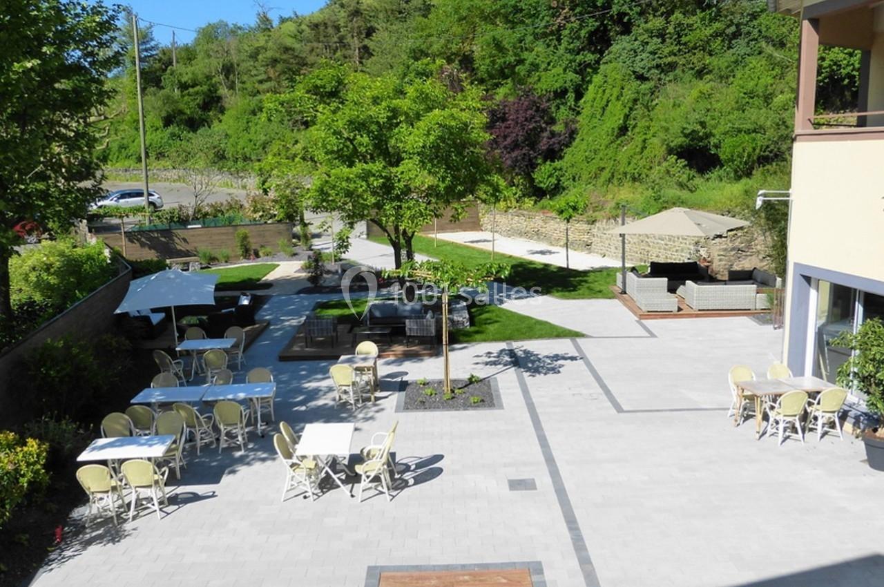 Terrasse extérieure avec tables et chaises, entourée de verdure et d'arbres, près d'un bâtiment jaune.