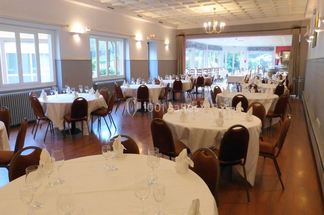 Salle de réception lumineuse avec tables rondes dressées, chaises en bois et grandes fenêtres donnant sur l'extérieur.
