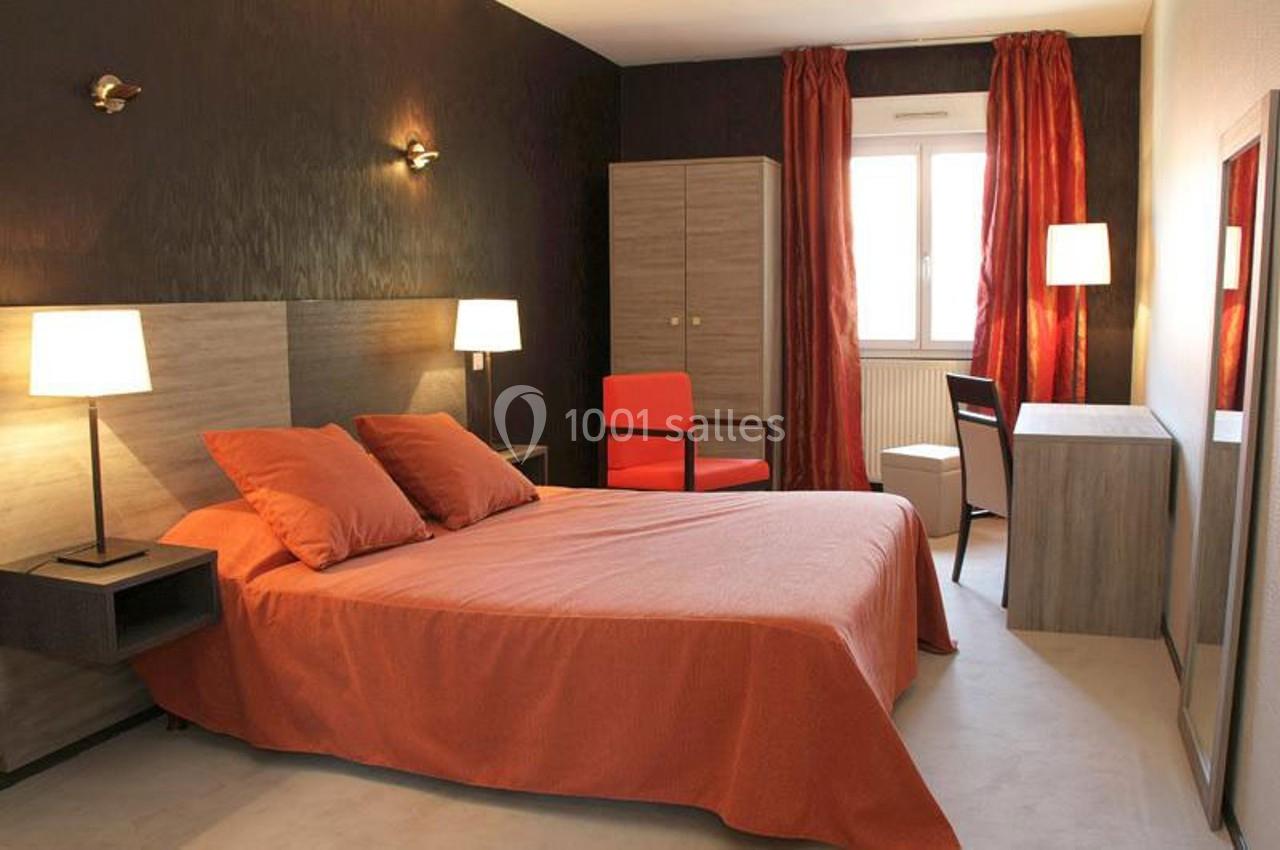 Chambre avec lit double, draps orange, bureau, chaise, fauteuil et rideaux assortis, éclairée par une fenêtre et des lampes.