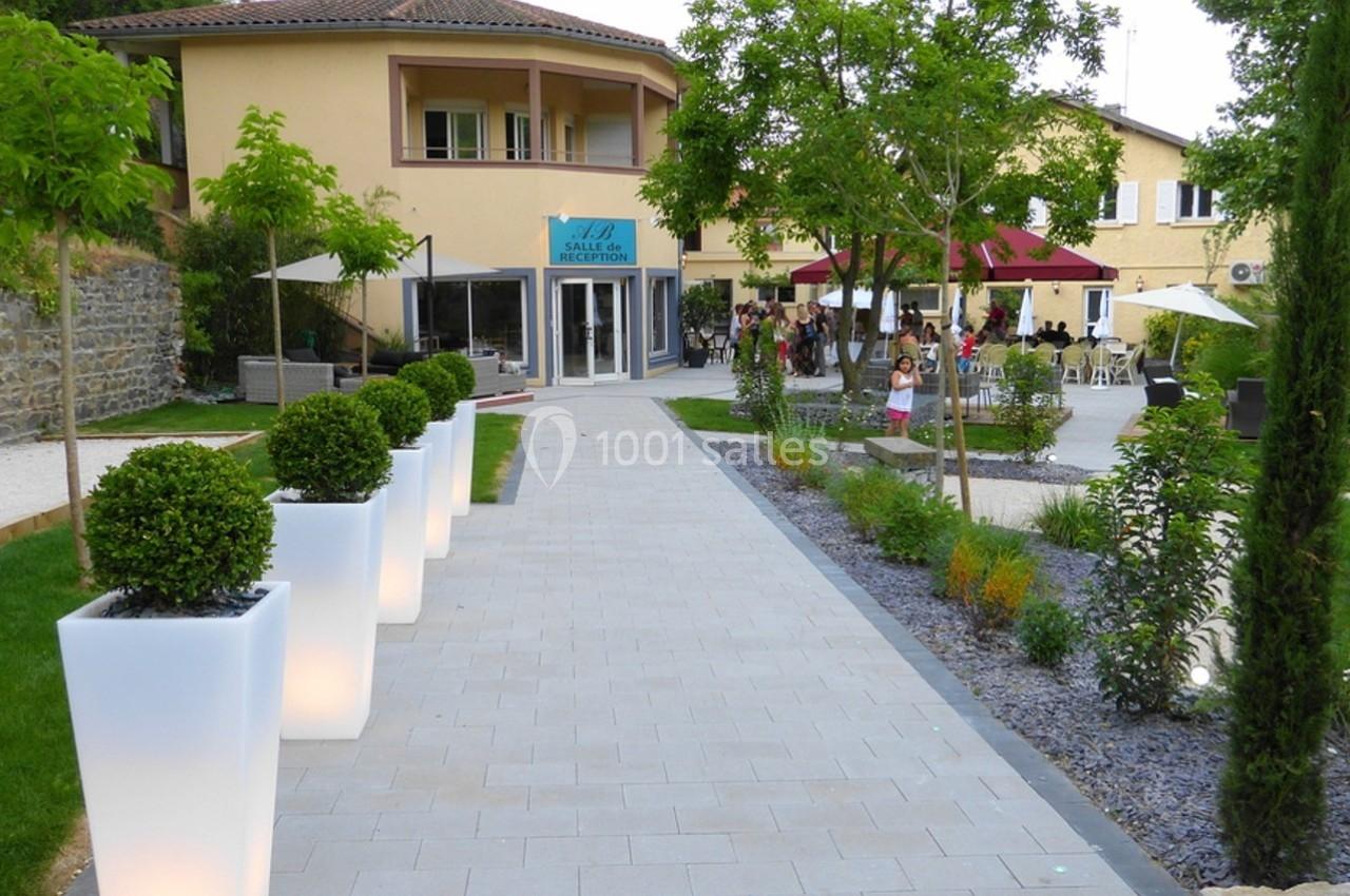 Allée pavée bordée de pots lumineux menant à un bâtiment avec terrasse et espaces verts aménagés.