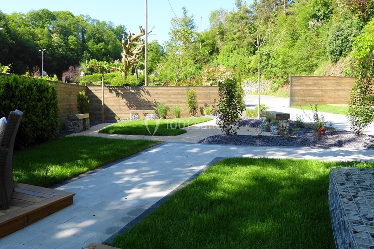 Jardin aménagé avec pelouse, allées pavées, parterres de plantes et clôtures en bois, entouré de verdure.