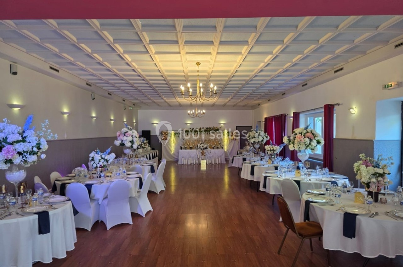 Salle de réception décorée pour un mariage, avec tables rondes, nappes blanches, fleurs et éclairage chaleureux.