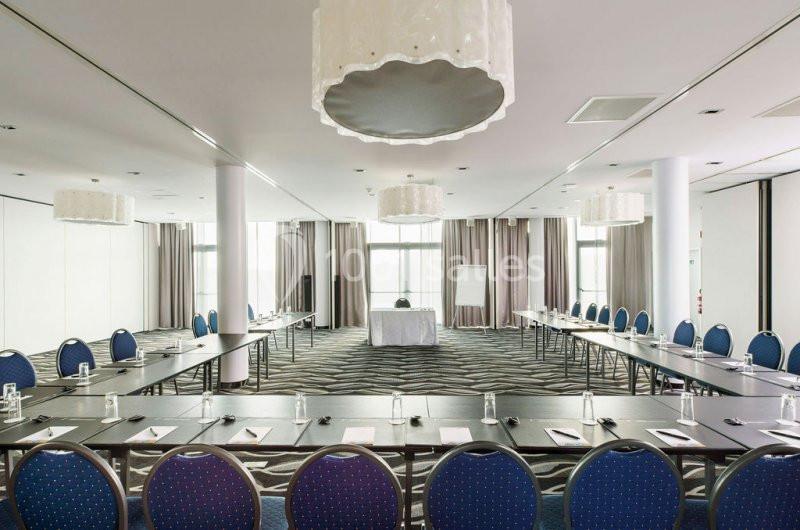 Salle de réunion lumineuse avec des tables disposées en U, des chaises bleues et un tableau de présentation.