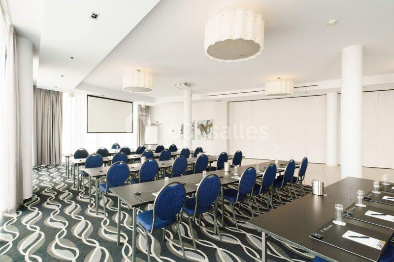 Salle de conférence lumineuse avec tables noires, chaises bleues, écran de projection et décor moderne.