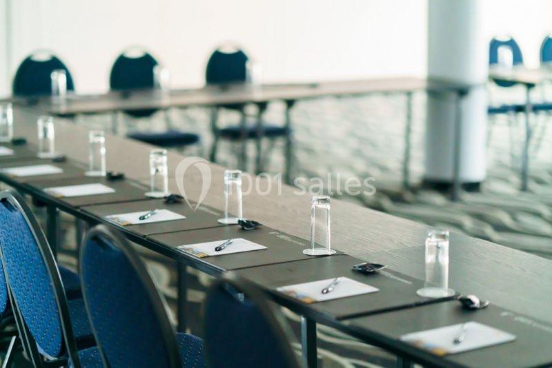 Salle de réunion avec des tables alignées, des verres, des stylos et des blocs-notes disposés devant des chaises bleues.