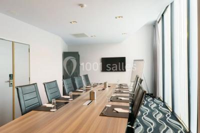 Salle de réunion moderne avec table rectangulaire, chaises noires, écran mural, tableau blanc et minibar.