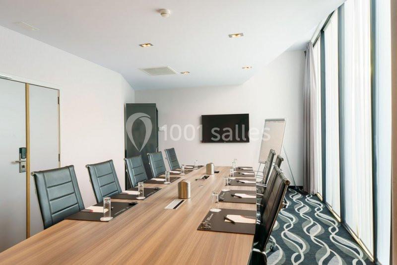 Salle de réunion moderne avec une grande table en bois, des chaises noires, un écran mural et un tableau blanc.