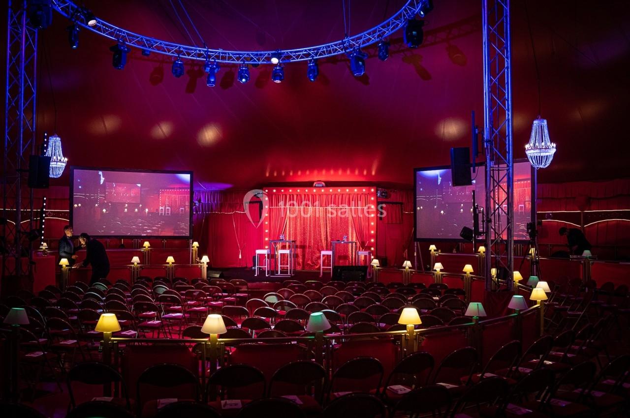 Intérieur d'un chapiteau éclairé, avec des rangées de chaises, des écrans et une scène décorée de rideaux rouges.