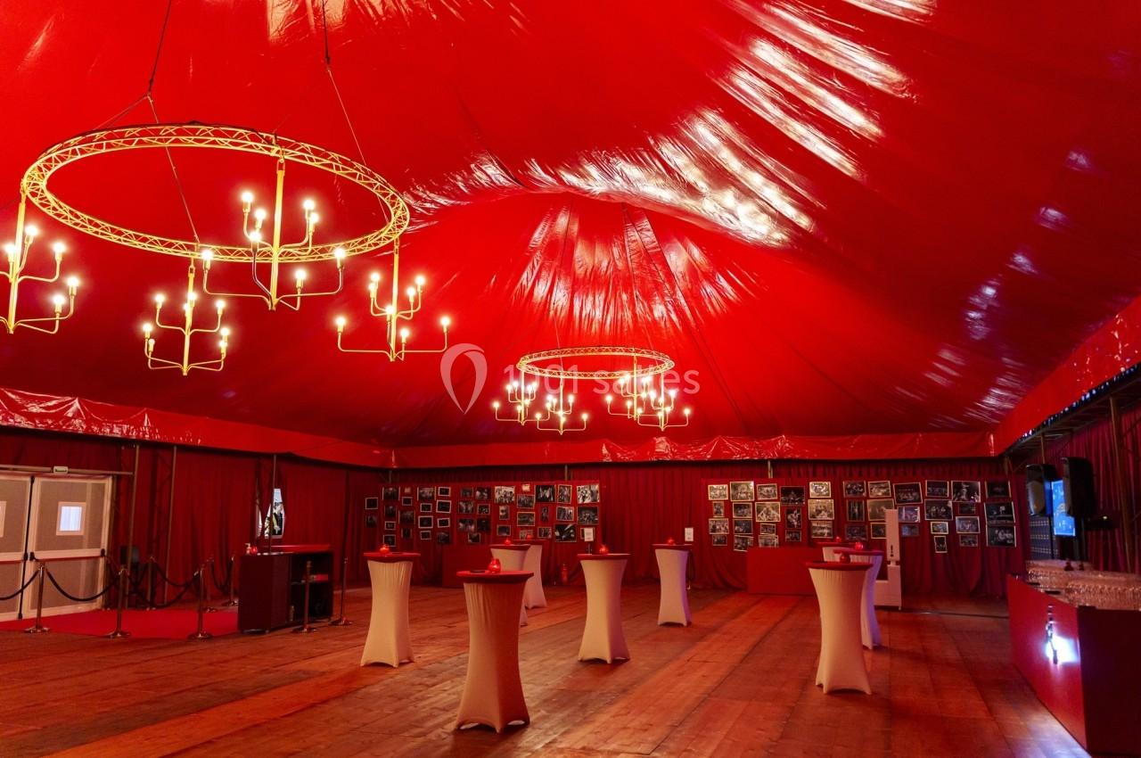 Salle sous chapiteau rouge avec lustres suspendus, tables hautes blanches et murs décorés de photos encadrées.