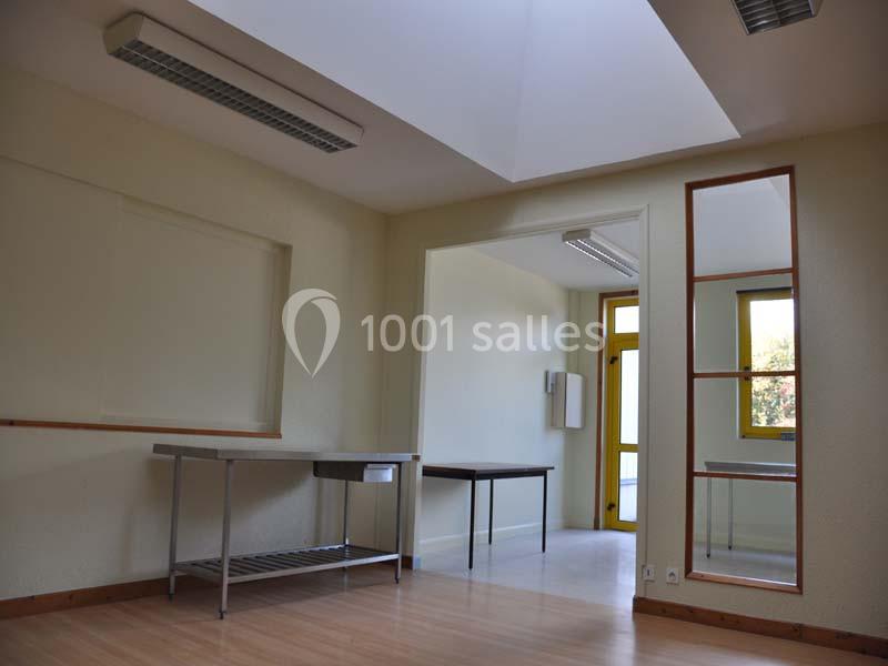 Salle vide avec parquet, murs clairs, table en métal, miroir mural et lumière naturelle entrant par une fenêtre.